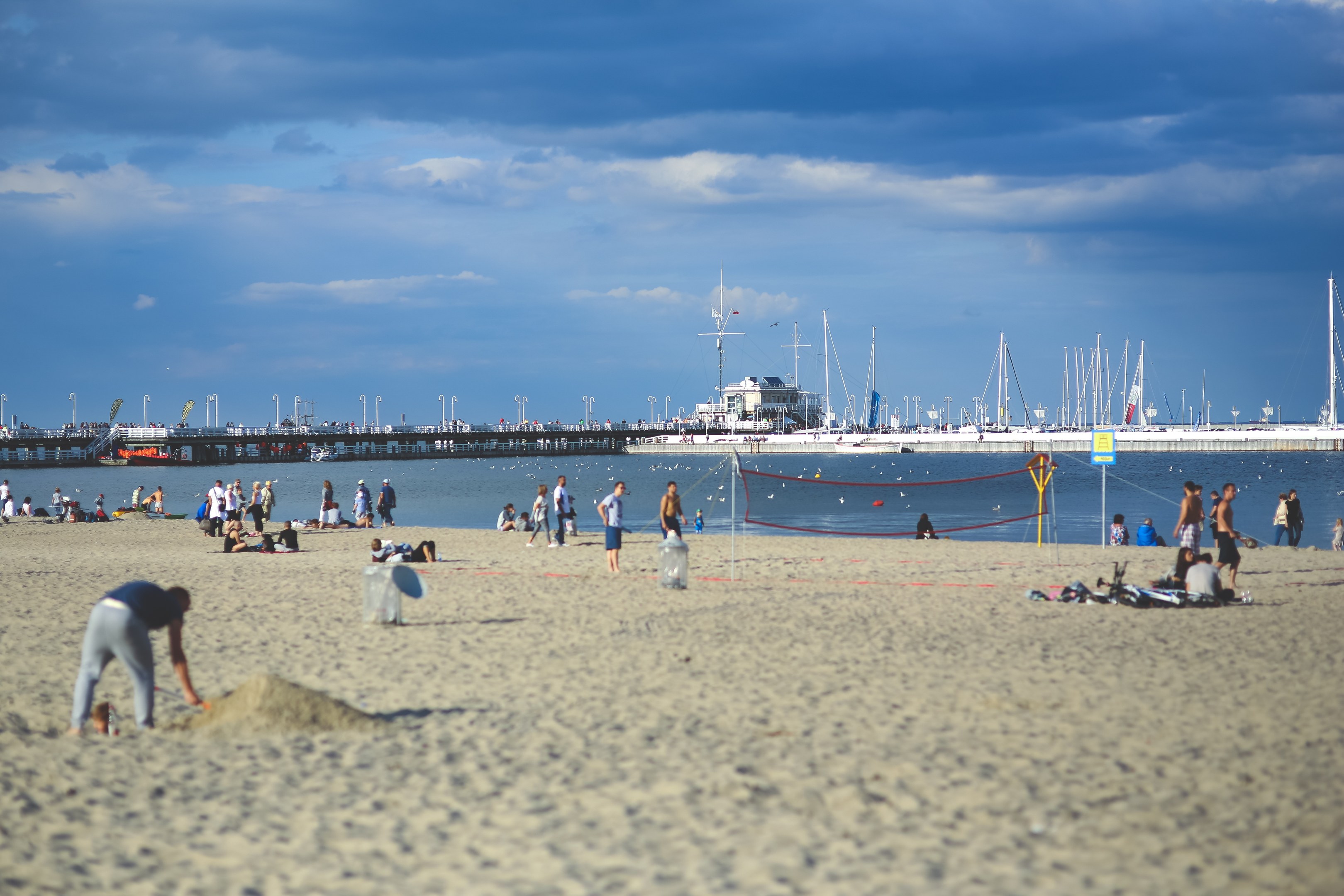 Menschen spielen Volleyball am Strand mit einem Netz im Sand, Boote und eine Brücke sind im Hintergrund unter einem bewölkten Himmel zu sehen.