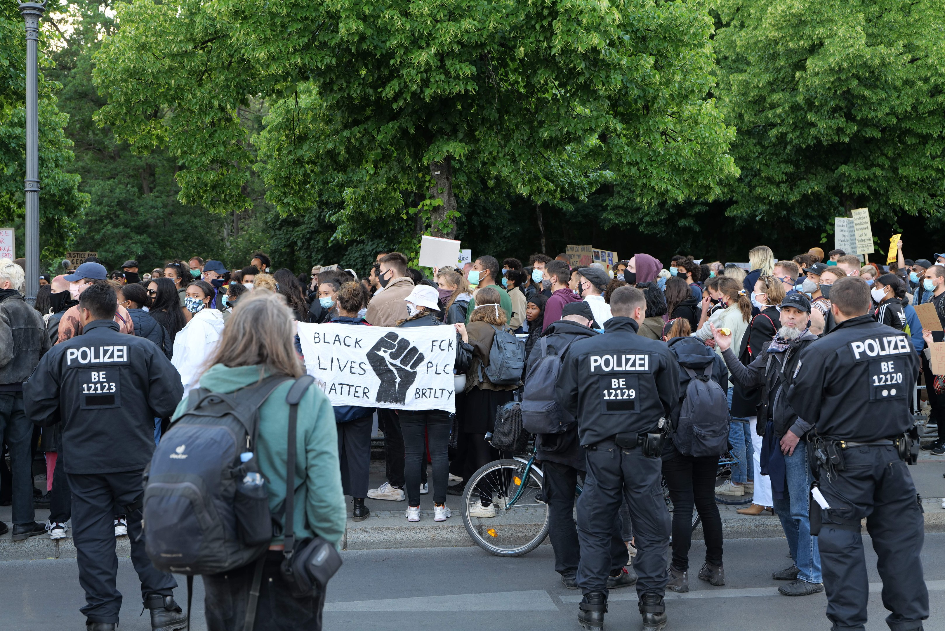 Eine große Gruppe von Menschen nimmt an einer Black Lives Matter Demonstration in Berlin teil, einige halten Schilder und andere tragen Mützen und Taschen, während im Vordergrund ein Fahrrad und im Hintergrund Bäume und ein Pfahl zu sehen sind.