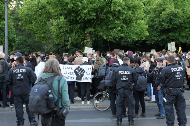 Eine große Gruppe von Menschen nimmt an einer Black Lives Matter Demonstration in Berlin teil, einige halten Schilder und andere tragen Mützen und Taschen, während im Vordergrund ein Fahrrad und im Hintergrund Bäume und ein Pfahl zu sehen sind.