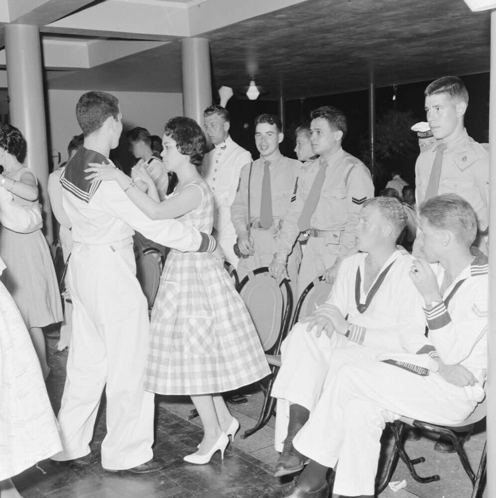 Schwarzes und weißes Bild von Militärpersonal, das auf einer Tanzfläche tanzt und sich vergnügt, mit Säulen und Beleuchtung im Hintergrund.