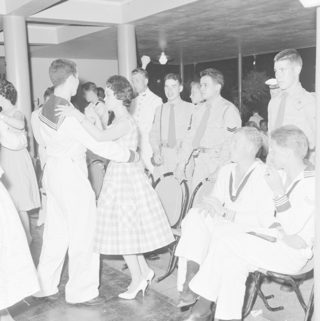 Schwarzes und weißes Bild von Militärpersonal, das auf einer Tanzfläche tanzt und sich vergnügt, mit Säulen und Beleuchtung im Hintergrund.