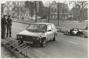 Ein Schwarz-Weiß-Foto von einem Auto, das auf der Straßenseite in ein Motorrad gekracht ist, mit drei Personen auf dem Gehweg links und Bäumen, Masten, Gebäuden und anderen Fahrzeugen im Hintergrund.