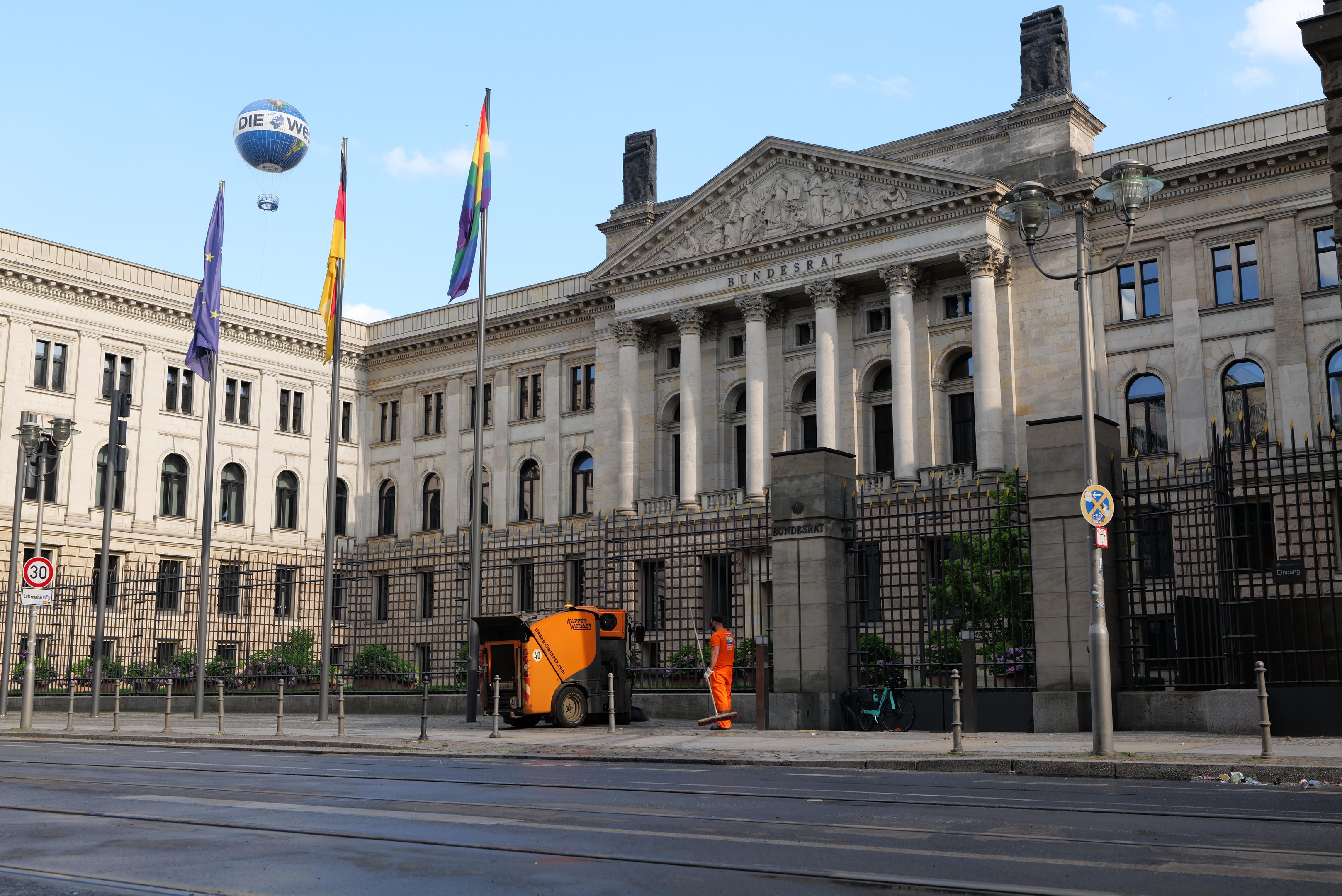 Großes modernes Regierungsgebäude mit vielen Fenstern, umgeben von Straßenmöbeln, Fahrzeugen, Fußgängern, Flaggen und Bäumen unter einem bewölkten Himmel.