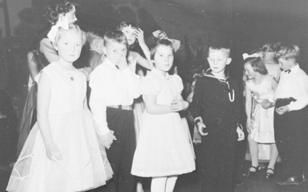 Schwarzes und weißes Bild einer Gruppe von Kindern, die auf einem Tanzboden stehen, lächeln und glücklich aussehen, mit einer Wand im Hintergrund.