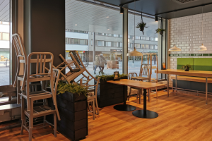 Ein Restaurant in Berlin, Deutschland, mit hölzernen Tischen und Stühlen in der Nähe eines großen Fensters, das Menschen auf der Straße, Gebäude und den Himmel zeigt, umgeben von Pflanzen und Lichtern.