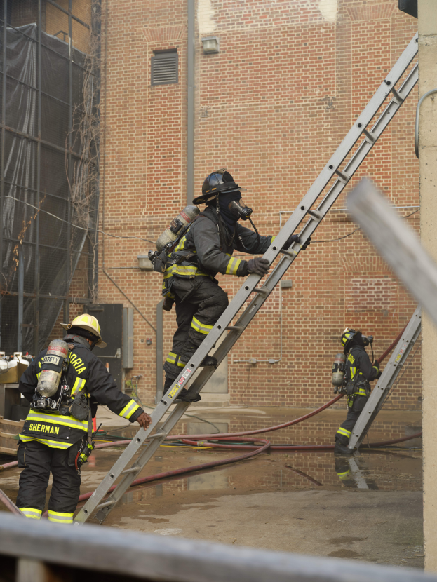Feuerwehrleute in Helmen und Zylindern klettern an einer Leiter vor einem Backsteingebäude hoch, mit Rohren und einem Metallstab auf dem Boden und einem Gebäude mit Fenstern und einem Netz im Hintergrund.