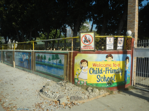 Ein Schultafel-Schild mit der Aufschrift "Willkommen in einer kinderfreundlichen Schule" neben einem Zaun, Bäumen, einem Weg und einem bewölkten Himmel.