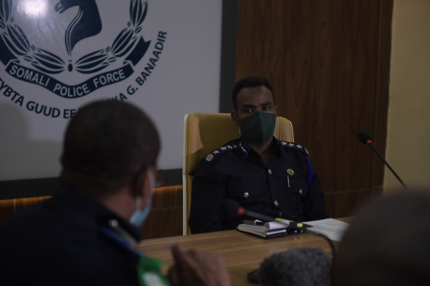 Zwei maskierte Polizeibeamte sitzen an einem Tisch mit Mikrofonen, Büchern und anderen Gegenständen und schauen auf einen Wandbildschirm mit Text und einem Emblem.