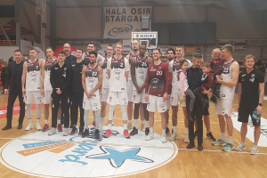 Gruppe von Männern und Kindern auf einem Basketballfeld mit Schildern "Hala Osir Starga", sitzende Zuschauer, ein Basketballkorb, Metallstangen, Deckenleuchten und eine Wand.
