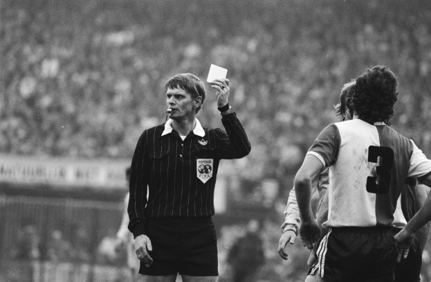 Schwarzes und weißes Bild eines Schiedsrichters, der eine rote Karte in der Luft hält, vor einer Menge von Menschen in Fußballtrikots, mit einem Zaun im Hintergrund.