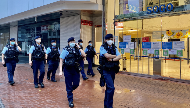 Eine Gruppe von Polizisten in Gesichtsmasken, die vor einem Glasgebäude die Straße entlanggehen, mit einem Geschäft auf der rechten Seite, das Poster an seinen Glaswänden hat.