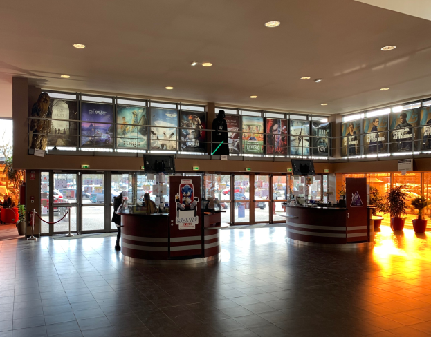 Lobby eines Kinos mit Menschen, Zimmerpflanzen, Fernsehern, Informationsschildern, Deckenleuchten, Glastüren und einer Außenansicht von Fahrzeugen, Bäumen, Gebäuden und Himmel.
