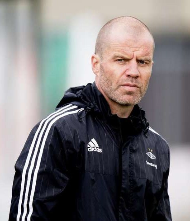 Kahlköpfiger Trainer in marineblauer Jacke mit weißen Streifen und weißem Kragen auf einem Fußballfeld stehend, die Hände gefaltet und einen ernsten Gesichtsausdruck.