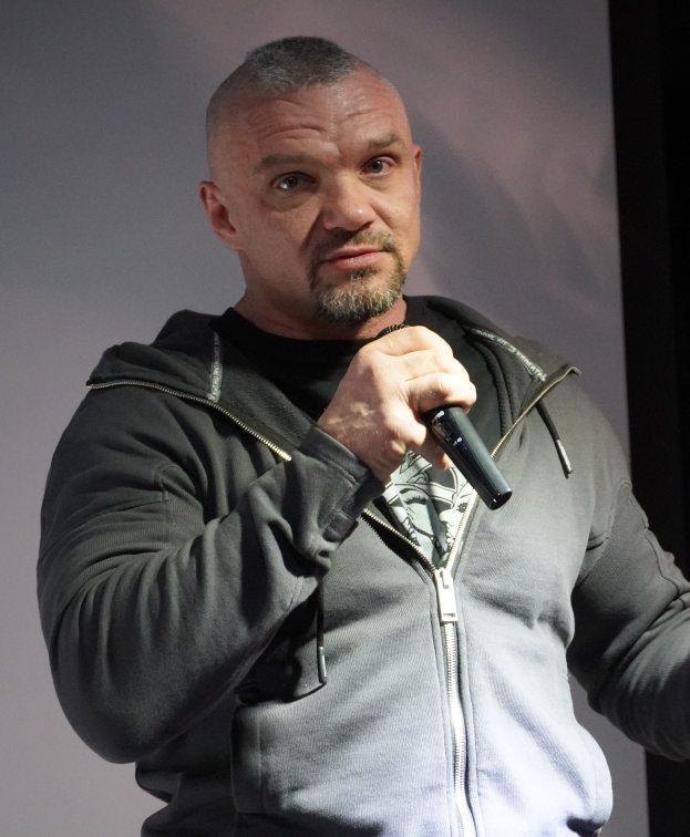 Comedian in a black suit and white shirt with slicked-back hair holding a microphone, standing confidently in front of a wall.