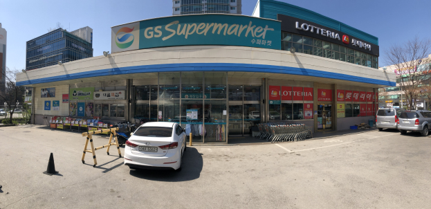 Ein Auto vor einem "GS Supermarkt"-Geschäft mit anderen Fahrzeugen, Verkehrskegeln und verstreuten Gegenständen auf der Straße, Gebäude und Bäume im Hintergrund bei klarem blauem Himmel.