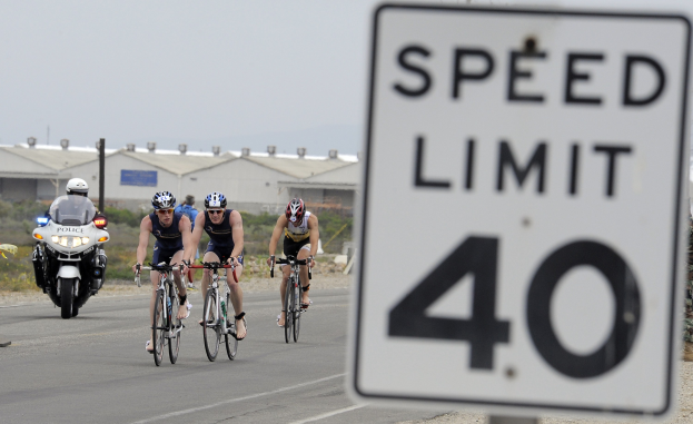 Gruppe von Fahrradfahrern in Helmen auf einer Straße neben einem Geschwindigkeitsbegrenzungsschild, mit Gebäuden, Versorgungsmästen, Vegetation und einem klaren blauen Himmel im Hintergrund.