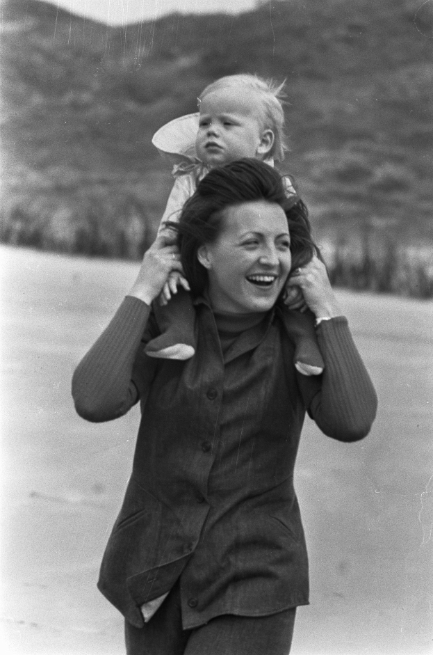Eine Frau mit warmem Lächeln hält ein Baby im Arm vor einem leicht unscharfen Hintergrund.