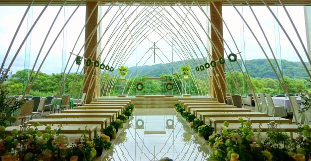 Eine Hochzeitszeremonie in einem großen Saal mit Tischen in Halbkreisform, geschmückt mit Blumen und anderen Gegenständen, vor einer Kulisse aus Bäumen, Hügeln und einem klaren Himmel.