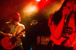 Eine Frau singt in ein Mikrofon auf der Bühne, während ein Mann Gitarre spielt und Deckenlampen die Szene beleuchten.