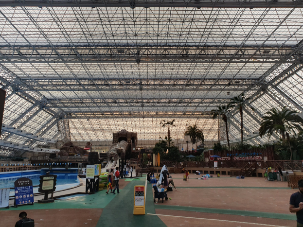 Gruppe von Menschen in einem Sommerland-Freizeitpark, die um ein Hallenbad mit Bäumen, Informationsschildern und einem klaren blauen Himmel im Hintergrund stehen und sitzen.