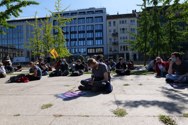 Eine Gruppe von Menschen sitzt vor einem Gebäude auf dem Boden während einer Demonstration in Berlin, einige tragen Masken, mit verstreuten Taschen und Gegenständen, unter einem klaren blauen Himmel mit Bäumen und Gebäuden im Hintergrund.