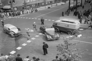 Eine Schwarz-Weiß-Szene eines Autounfalls an einer Straßenkreuzung mit mehreren Fahrzeugen, darunter einem Van, einer Gruppe von Menschen drumherum und im Hintergrund Gebäuden, Bäumen, Pfählen und Texttafeln.