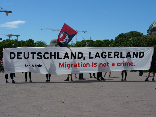 Eine Gruppe von Menschen mit einem Transparent mit der Aufschrift "Deutschland, Lagerland Migration ist kein Verbrechen" und einer Flagge, im Hintergrund sind Laternenpfähle, Lichter, Bäume, Kräne, ein Gebäude und ein bewölkter Himmel zu sehen.