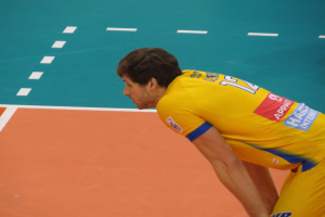 Ein professioneller Volleyballspieler in einem gelben "Volleyball"-T-Shirt steht selbstbewusst auf einem Court und ist bereit, den Ball zu servieren.
