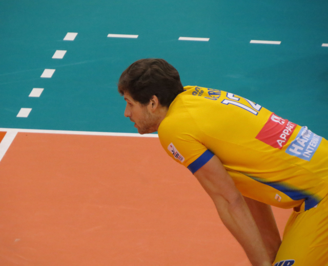 Ein professioneller Volleyballspieler in einem gelben "Volleyball"-T-Shirt steht selbstbewusst auf einem Court und ist bereit, den Ball zu servieren.
