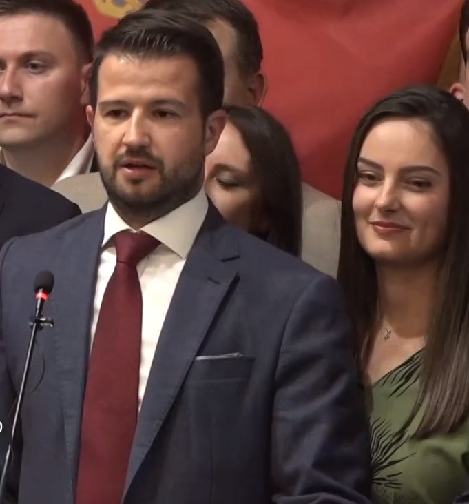 Ein Mann im Anzug spricht an einem Podium mit einem Mikrofon vor einer Menge, während im Hintergrund ein Banner hängt.