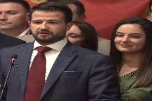 Ein Mann im Anzug spricht an einem Podium mit einem Mikrofon vor einer Menge, während im Hintergrund ein Banner hängt.