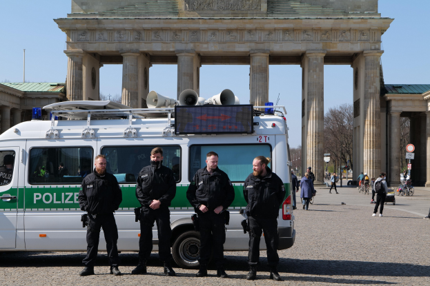 Polizisten in schwarzen Uniformen stehen vor dem Brandenburger Tor in Berlin, mit einem weißen und grünen Fahrzeug im Vordergrund und Passanten und Radfahrern im Hintergrund.