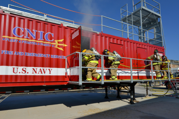 Eine Gruppe von Feuerwehrleuten in Helmen mit Rohren steht auf einem roten Seecontainer mit Text, Geländern und Stufen, mit einem Fahrzeug auf der Straße, Bäumen und einem bewölkten Himmel im Hintergrund.