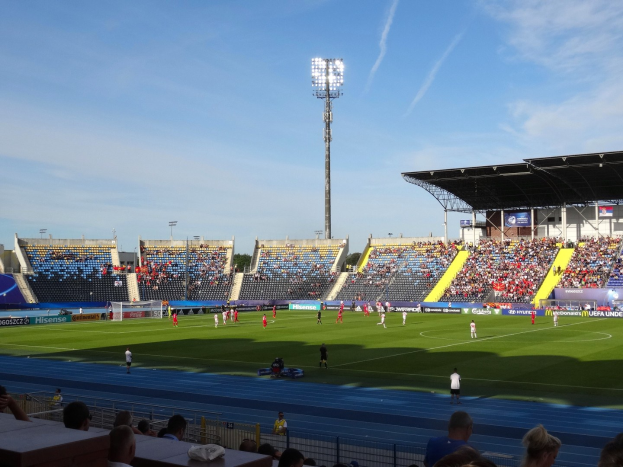 Ein Fußballspiel im Gange in einem großen, beleuchteten Stadion mit Zuschauern auf den Rängen und Spielern auf dem Feld unter einem sichtbaren Himmel.