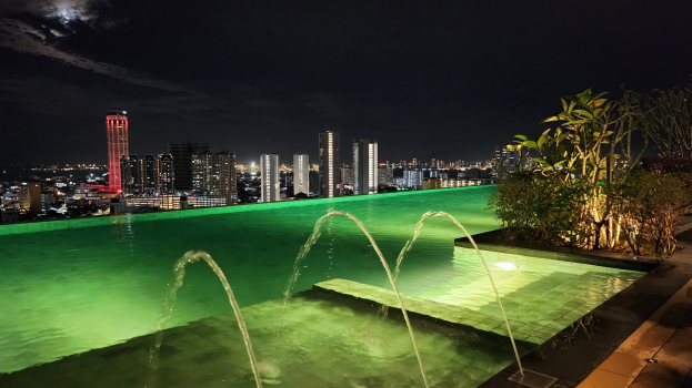 Ein Schwimmbad mit einem zentralen Brunnen, umgeben von üppiger Vegetation auf der rechten Seite und von Gebäuden, die von Stadtlichtern im Hintergrund beleuchtet werden.