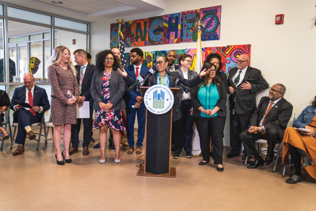 Gruppe von Menschen vor einem Rednerpult mit einem Mikrofon und einer Tafel mit der Aufschrift "Sancho-Stadtrat tritt der Stadtvertretung bei", mit sitzenden und stehenden Teilnehmern, Flaggen, einem Feuerlöscher und Deckenleuchten im Hintergrund.