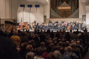 Eine große Gruppe von Menschen sitzt vor einer Bühne bei einem Konzert, einige halten Musikinstrumente, Fahnen, eine Lautsprecherbox und andere Gegenstände sind auf der Bühne zu sehen, es gibt eine Decke, Deckenleuchten und eine T├╝r an der hinteren Wand.