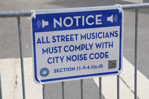 Anschlagtafel an einem Zaun mit einem Schild, das die Einhaltung der Lärmvorschriften für Straßenmusiker vorschreibt, mit einer Straße im Hintergrund.