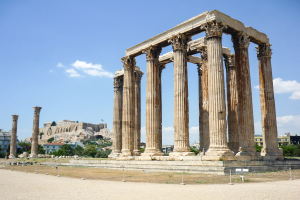 Alter Tempel von Olympian Zeus in Athen, Griechenland, mit hohen korinthischen Säulen, umgeben von Bäumen und Felsen, mit einer Burg und einem bewölkten Himmel im Hintergrund.