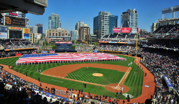 Baseballstadion mit Zuschauern, einer amerikanischen Flagge über einer Gruppe in der Mitte, Anzeigetafeln an den Seiten des Feldes und Gebäuden, Bäumen und einem blauen Himmel im Hintergrund.