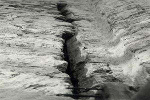 Schwarzes und weißes Foto eines große Riss im Boden, wahrscheinlich ein Entwässerungskanal, mit einem sichtbaren Wasserzeichen auf der linken Seite.