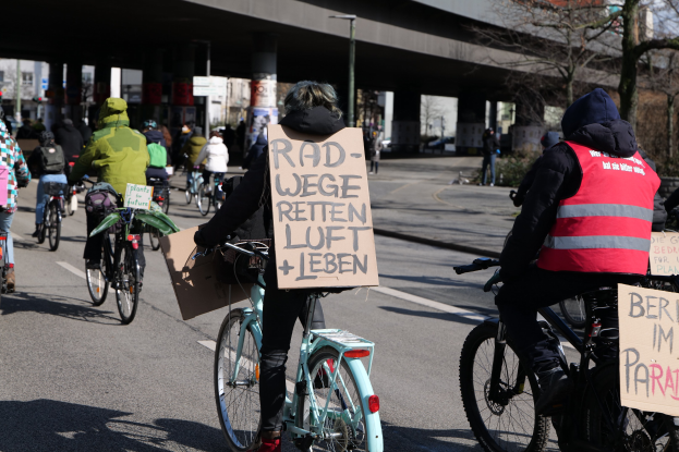 Gruppe von Menschen, die auf Fahrrädern eine Straße mit einer Brücke im Hintergrund entlangfahren, mit Bäumen an der Straße und Gebäuden in der Ferne, die Schilder mit verschiedenen Botschaften halten.