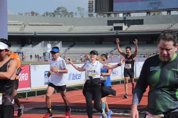 Gruppe von Menschen, die auf einer Outdoor-Sportanlage laufen, mit Bannern, einer Anzeigetafel, einem Gebäude, Bäumen und einer bewölkten Himmel; einige Läufer tragen Turnschuhe und Hüte.