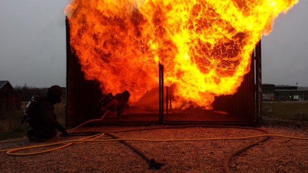 Feuerwehrleute löschen ein Gebäude Feuer, während ein Rohr auf dem Boden liegt, mit Schuppen, Bäumen und einem blauen Himmel im Hintergrund.