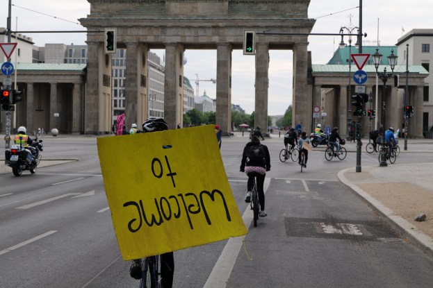 Eine Gruppe von Radfahrern fährt an der Brandenburgertor in Berlin vorbei, einer hält ein gelbes Schild, mit Laternenmasten, Verkehrsampeln, Gebäuden, Bäumen und einem klaren Himmel im Hintergrund.