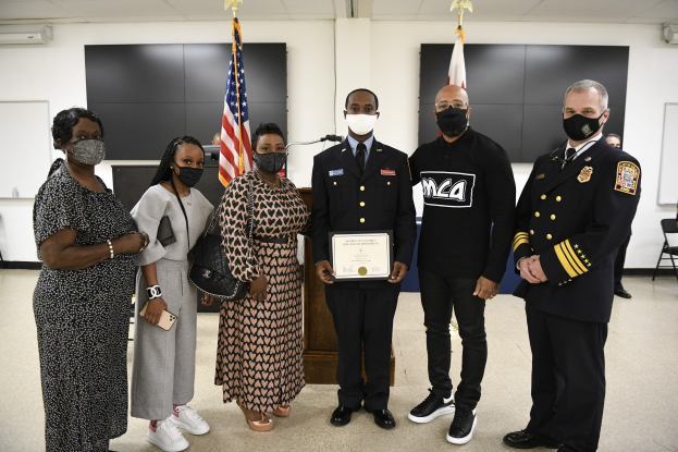 Eine Gruppe von Menschen in Masken steht zusammen vor Flaggen, hält ein Zertifikat, wahrscheinlich bei einer Abschlussfeier, mit Stühlen, Tischen und anderen Gegenständen im Hintergrund.