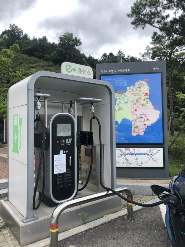Elektrofahrzeug-Ladestation mit einem digitalen Display, das anzeigt, dass es eine E-Bike-Ladestation ist, ein Motorrad in der Nähe geparkt, Bäume und klarer blauer Himmel im Hintergrund.