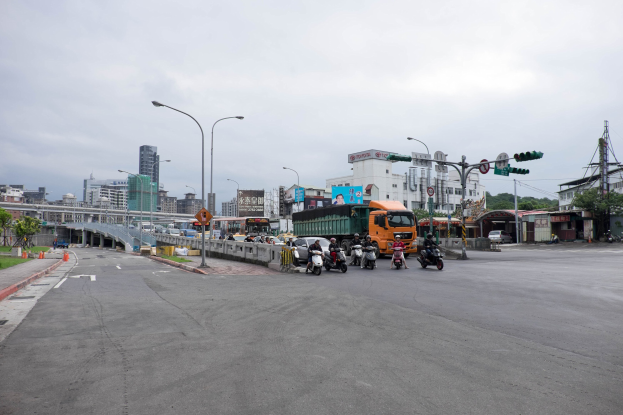 Stadtstraße mit starkem Verkehr, einschließlich eines Lkw und eines Autos, mit Laternen, Verkehrsampeln, Schildern, einer Brücke mit Geländer, Bäumen, Gebäuden mit Fenstern und einem bewölkten Himmel.