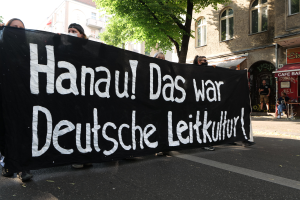 Eine Gruppe von Menschen, die eine schwarze Fahne mit der Aufschrift "Hanau das war deutsche Leitkultur" tragen, geht öffentlich spazieren, mit Gebäuden, Bäumen und einem klaren blauen Himmel im Hintergrund.
