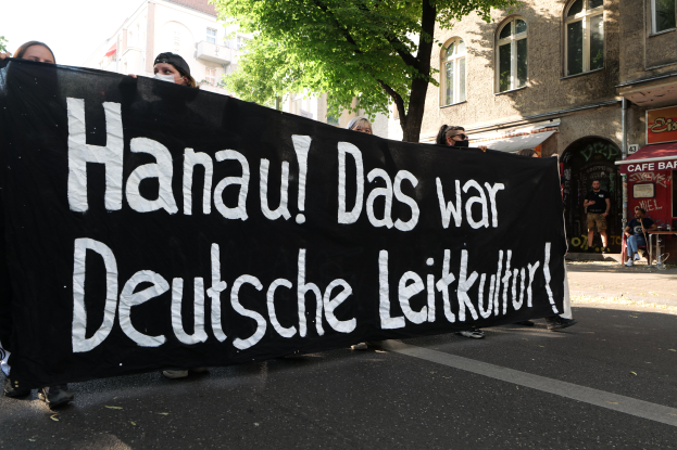 Eine Gruppe von Menschen, die eine schwarze Fahne mit der Aufschrift "Hanau das war deutsche Leitkultur" tragen, geht öffentlich spazieren, mit Gebäuden, Bäumen und einem klaren blauen Himmel im Hintergrund.